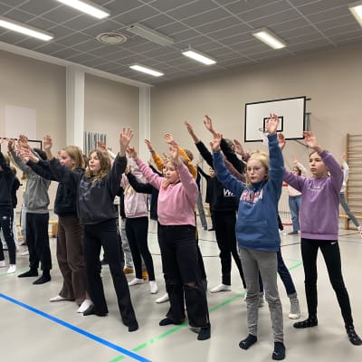 Iso lapsiryhmä seisoo kädet ylhäällä koulun liikuntasalissa. Hannulan koulun oppilaat ovat harjoittelemassa koululaisoopperaa.