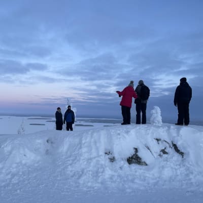 Ihmiset valokuvaavat Ukko-Kolin huipulla talvisen illan sinisessä hämärässä.