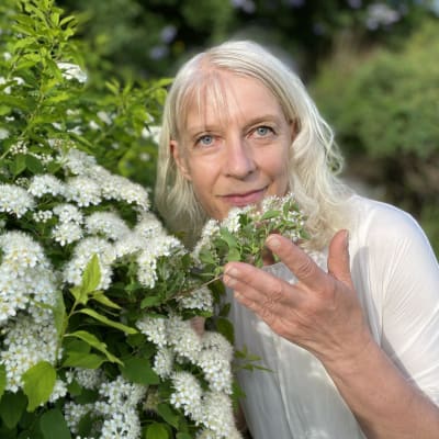 Vaalea nainen (Aija Matikainen) nostaa yhtä valkoista kukkaterttua pensaasta kohti kameraa ja katsoo kameraan päin. 