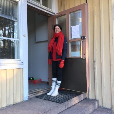 Nainen pitää vanhan kyläkoulun ovea auki nojaamalla siihen.