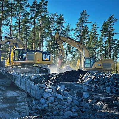Två grävmaskiner på larvfötter arbetar bland krossgrus och stenbumlingar men att jämna marken. 