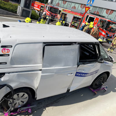 Palanut ja sammutettu valkoinen sähköauto hinauksessa parkkihallista tielle. Taustalla useita palomiehiä ja kaksi paloautoa.