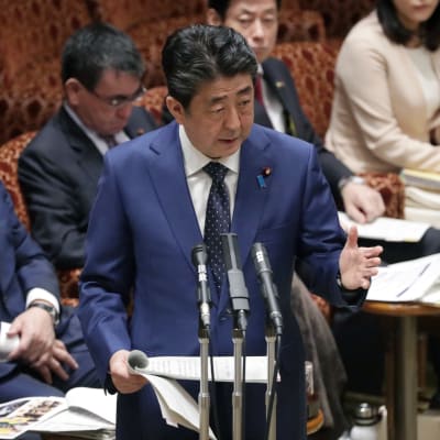 Japans premiärminister Shinzo Abe står i ett talarpodie i Japans parlamet och håller tal om bland annat att sommar-OS i Tokyo kan komma att flyttas p.g.a coronapandemin. I bakgrunden sitter fyra personer, varav en bär ansiktsmask.