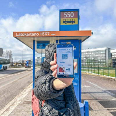 En person visar sin telefon, som visar en nätsida med en bussbiljett. I bakgrunden en busshållplats.