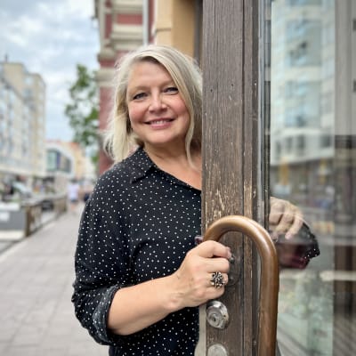 Riikka Haapasalo katsoo kameraan ja hymyilee avatun oven takaa. Hänellä on pitkät hiukset ja musta paita, jossa on valkoisia pilkkuja. 
