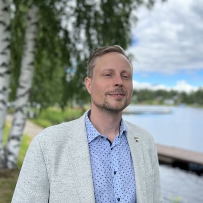Mies harmaassa pikkutakissa ja vaaleansinisessä paidassa seisoo veden äärellä.