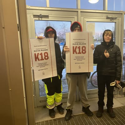 Kolme nuorta kantaa k18-kylttejä haminalaisen ABC:n tuulikaapissa.