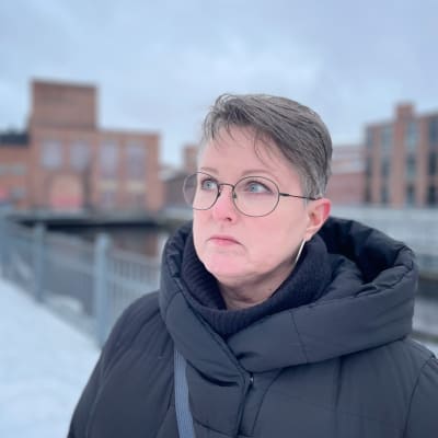 Näyttelijä ja laulaja Leena Rousti seisoo kosken rannalla, Museokeskus Vapriikin vieressä. Taustalla näkyy punatiilisiä rakennuksia. Leenalla lyhyet hiukset ja musta toppatakki. 