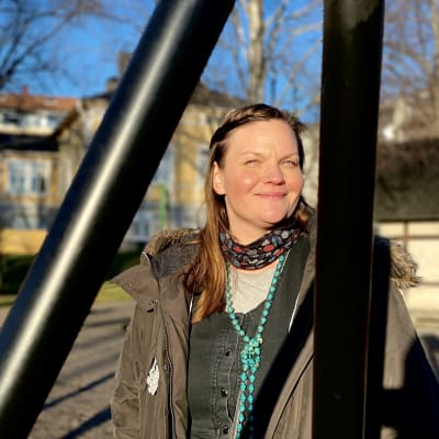 Nainen seisoo auringossa. Hänet rajaa pihakeinun jalat. Paikka on puisto.
