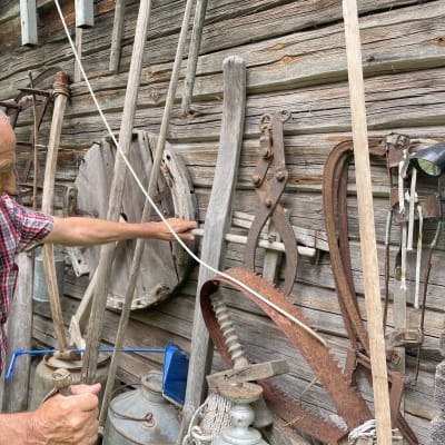 84-vuotias Erkki Kauppinen esittelee vanhoja puisia työkaluja, joita on ripustettu vanhan harmaan aitan seinälle.