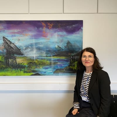 Aineen taidemuseon johtaja Virpi Kanniainen istuu Ville Rädyn maalauksen vierellä. Maalauksessa muun muassa pieni joki, maastoa ja taivasta.