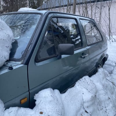 Vanha auto, jonka keulan ja etulasin päällä on lunta. Myös auton ympärillä on kinos.