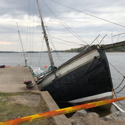 En stor båt med mast, kanske trålare som håller på att sjunka i vatten, nära strand.