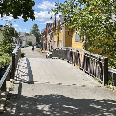 Katu, jonka varrella on keltaisia puisia taloja, joissa on harmaa harjakatto. Etualalla on kaareva kävelysilta, jossa on harmaat rautakaiteet. Alla virtaa vettä. Aurinko valaisee katua, pyöräilijä kulkee sillä kauempana. 