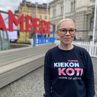 Tampereen kaupungin tapahtumapäällikkö Saara Saarteinen päällään "Kiekon koti" -tekstillä varustettu tummansininen paita. Taustalla iso "Tampere"-kyltti.
