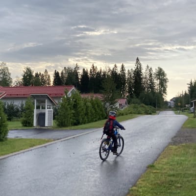 Poika ajaa polkupyörällä reppu selässään katua pitkin.