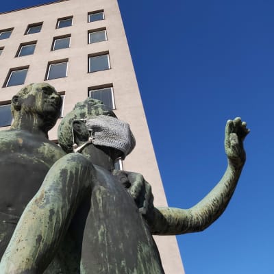 Kuva Kotkan kaupungintalolta patsaasta, jossa on mies ja lapsi. Lapsipatsaan kasvoilla on neulottu kasvomaski.