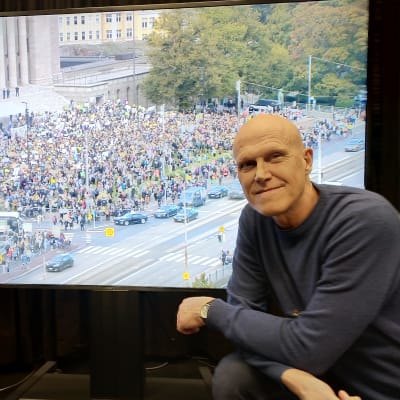 Thomas Wallgren i Svenska Yles lokaler framför en tv-skärm. På tv-bilden syns klimatdemonstranter utanför riksdagen den 27 september.