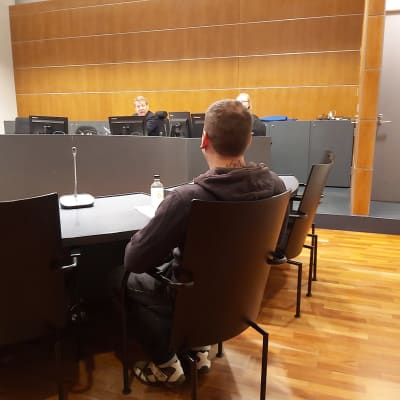 En ung man sitter på en stol i en rättssal, ryggen mot betraktaren. Han står åtalad för grovt skattebegrägeri i vad som är Finlands första Onecoin-rättegång.