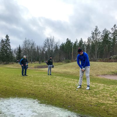 Tre män står på en golfbana, en puttar medan de andra ser på.