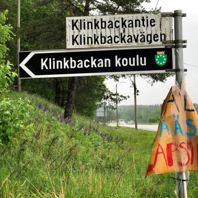 En vägskylt med finsk och svensk text. Nedanför vägnamnen står det på en svartvit skylt Klinkbackan koulu. Till höger löper en asfaltväg. Regnig väder. Grön försommarmiljö.