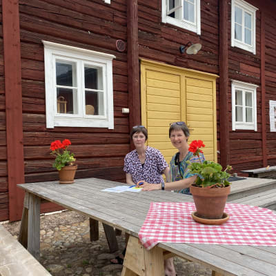 Susanne Lagus och Gunilla Sand sitter utomhus vid ett bord med papper framför sig på bordet och en röd stockvägg bakom sig.