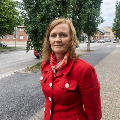 Kvinna i röd jacka står på trottoar. Bilar och träd i bakgrunden.