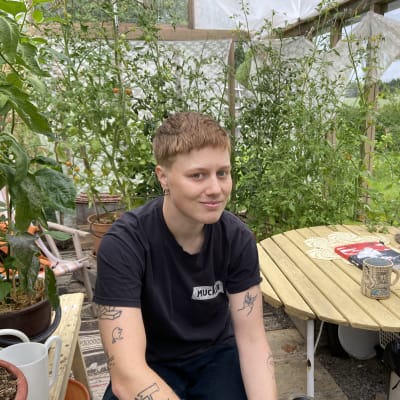 Edith Hammar sitter i sitt växthus och tittar in i kameran. I bakfgrunden skymtar växter och gröna kvistar, och boken "Homo Line" på bordet. 