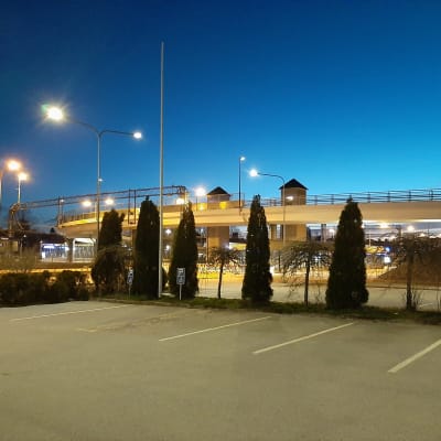 Asfalterade parkeringsplatser och gröna buskar framför en vit bro nattetid.