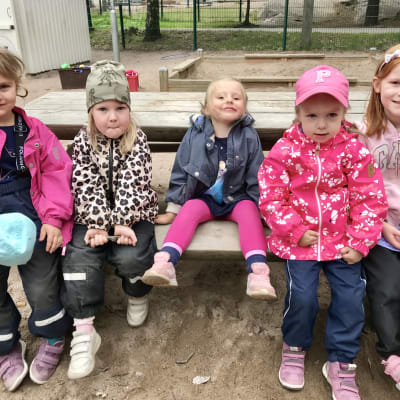 Fem flickor sitter på en bänk utomhus.