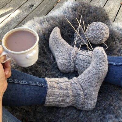 Fötter iklädda stickade sockor, stickning och garnnystan på fårfäll. En hand som håller en kopp varm choklad.