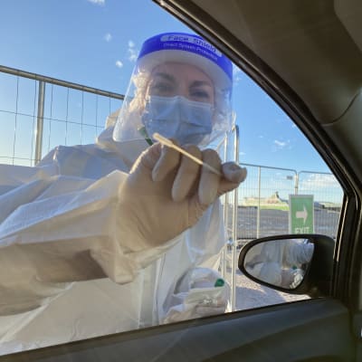 En person i munskydd och visir sträcker in en teststicka i en bil.
