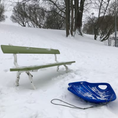 Parkbänk och pulka nedanför snöig backe.
