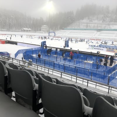 Hiihtäjiä ja mediaa kisamontussa sumuisena päivänä, katsomon penkit ovat tyhjinä 