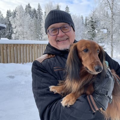 Mies koira sylissään talvisessa lumimaisemassa
