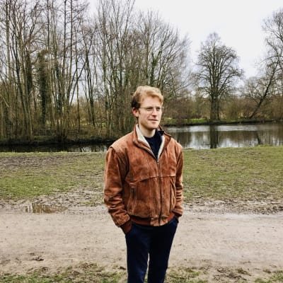 23-åriga studeranden Ollie Charlton i fotograferad i en park i Amsterdam. Han är ledigt klädd. Det är fortfarande tidig vår och inga löv i träden. 