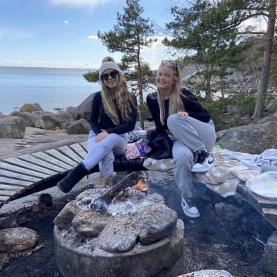 Pinja och Riina sitter och ler vid en öppen eld med havet i bakgrunden.