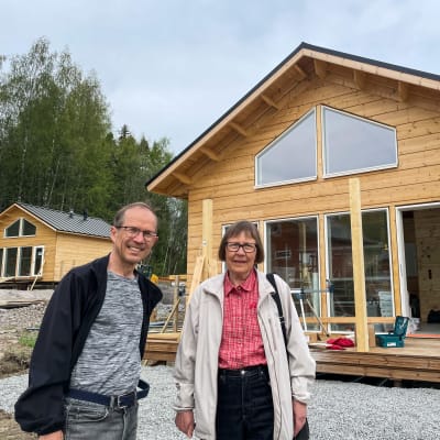 Pekka och Kristiina står framför sin sommarstuga i trä.