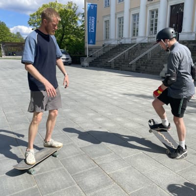 Peik Erhola opastaa pappi Olli Viljakaista rullalautailuun Tampereen vanhan kirjastotalon edustalla. 