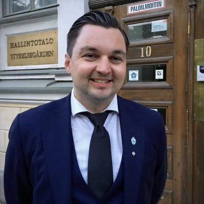 Man i blå kavaj står utanför en gul byggnad.