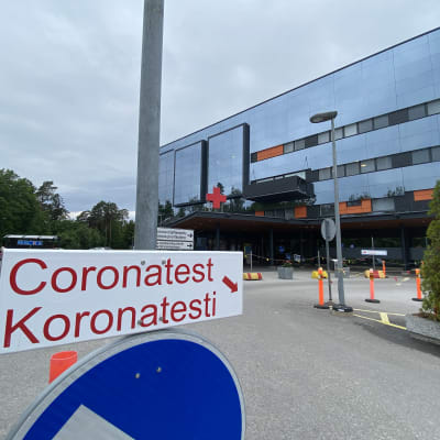 En skylt till corona provtagning. I bakgrunden ses en sjukhusbyggnad.