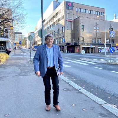 Kirjailija Timo Sandberg kävelee kadulla, taustalla kaupunkinäkymä ja kauppakeskus Trio Lahdessa.