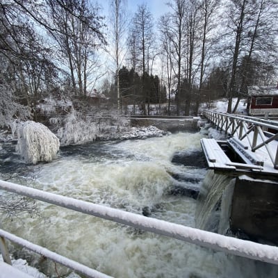 Kosken vesi kuohuu valkoisena ja padon yli rakennettu silta, johtaa punaiselle mökille. Puut ovat kuurassa pakkasesta.