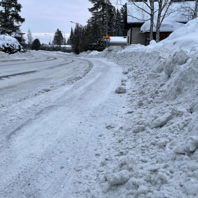 Tien pätkä, missä lunta kasautunut aurauksen myötä runsaasti jalkakäytävän reunaan sekä tielle.