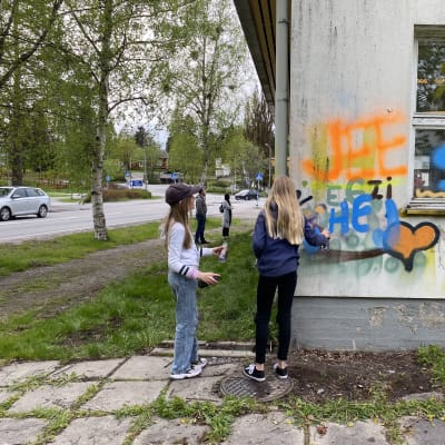 Två unga tjejer målar grafitti på skolbyggnad.