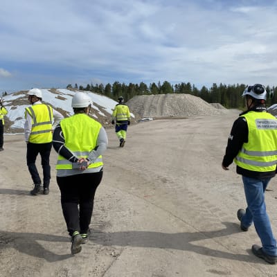Fem personer klädda i gula västar och vita hjälmar framför stora grushögar.