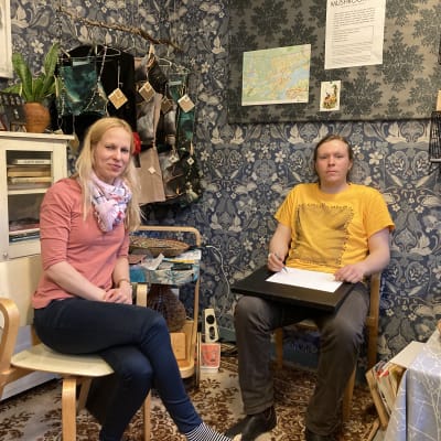 Nainen ja mies istuvat Tampereen Kauppahallissa näyttelytilan tuoleilla. Miehen vieressä pöydällä on pullavati ja naisen takana vaaterekki..