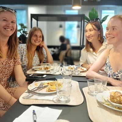 Fyra damer i sommarklänningar sitter i en studentmatsal med matbrickor framför sig.
