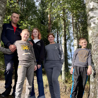 Äiti, isä ja kolme lasta poseeraavat seisten kameralle kahden koivun välissä.
