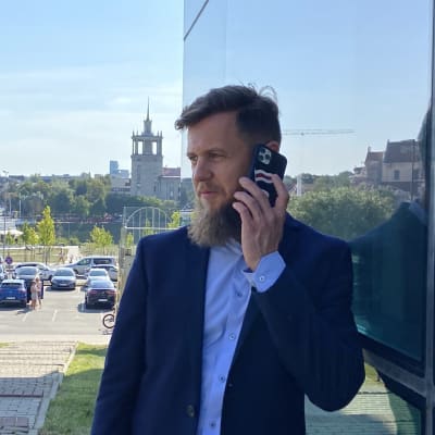 Man med blå kostym och ljusblå skjorta talar i mobiltelefon utanför ett konferenshotell i Vilnius. Mannens mobiltelefon pryds av Belarus rödvita oppositionsflagga.